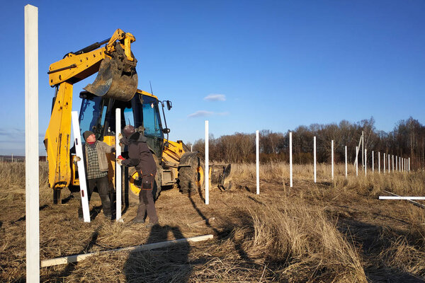The excavator with the bucket clogs the iron pillars for the fence in the field, the pillars painted with white paint 2019