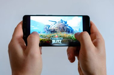 Los Angeles, California, USA - 9 March 2019: The man holds the smartphone close-up in his hands and plays the world of tanks blitz against the background of a colored blurred side. clipart