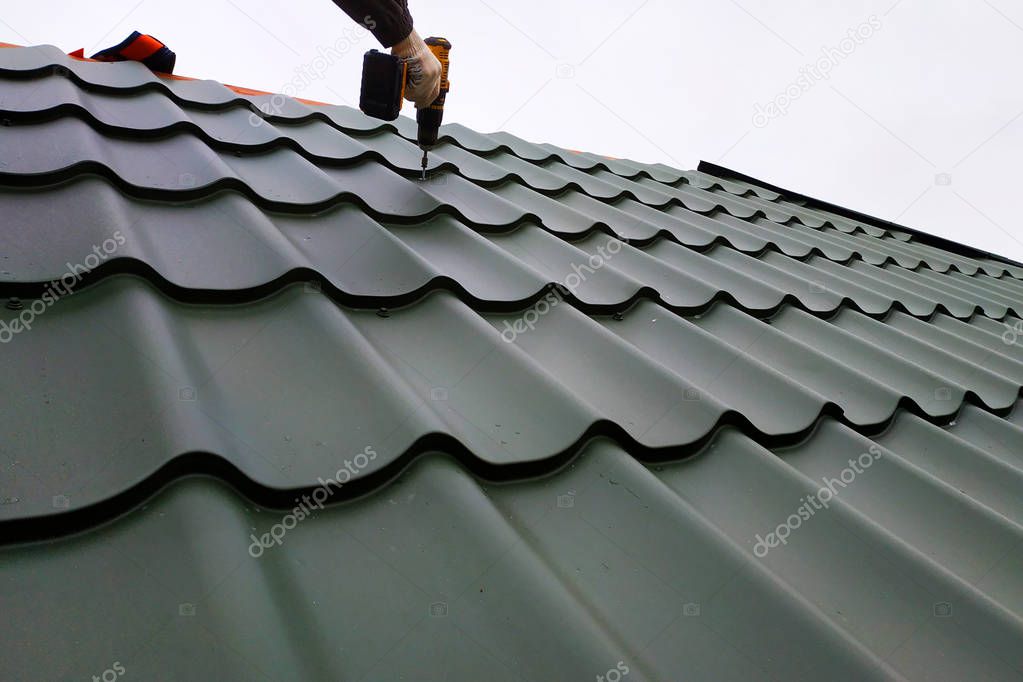 The professional worker works on installation of a roof of a roof by sheets of a metal tile and drills a screw with a drill