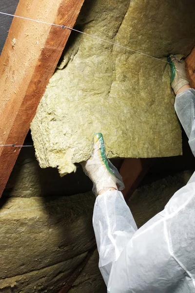 Un hombre con un mono protector pone lana mineral entre la viga del techo de la casa para su calentamiento del frío —  Fotos de Stock