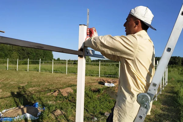 İşçi merdivende duruyor ve açısal cetvel çitin metal profilinin ölçümlerini kaldırır.