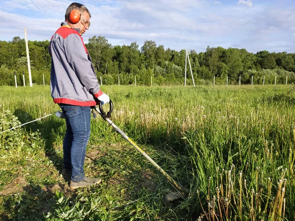 Um homem rema grama com um cortador de carvalho no campo, no quintal é verão e o sol brilha . — Fotografia de Stock