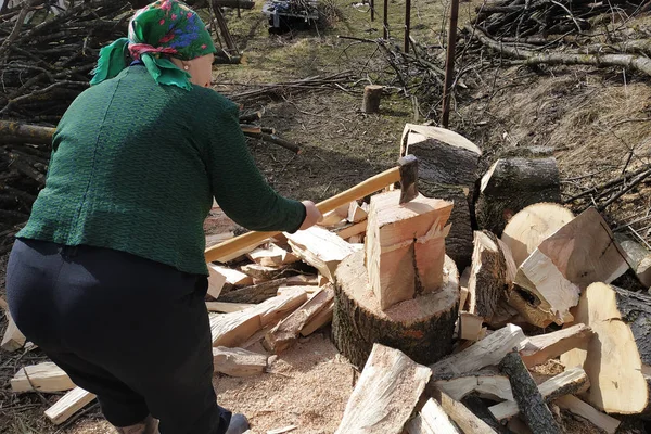 stock image An old woman of a tavern shakes a firewood with an ax in the yard, prepares them for the winter.