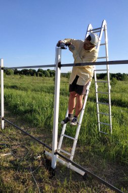 Bahçedeki bir adam çitin üzerindeki metal profili köşe öğütücüyle merdivende durur..