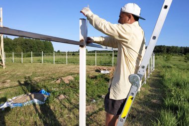 İşçi merdivende duruyor ve açısal cetvel çitin metal profilinin ölçümlerini kaldırır.