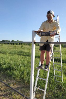 Bahçedeki bir adam çitin üzerindeki metal profili köşe öğütücüyle merdivende durur..
