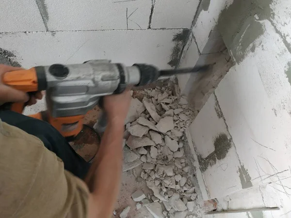 Un trabajador con un perforador rompe la pared y los cimientos de la casa para conducir cables eléctricos . — Foto de Stock