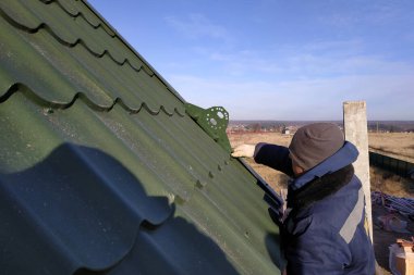 Soğuk sezonun ustası çatıda çalışıyor, karları parçalayan bir tornavidayla bağlanıyor, onları galvanize çatıya dikkatlice uyguluyor, çok dikkatli davranıyor, yükseklikte çalışıyor, yeni bir ev. 2020