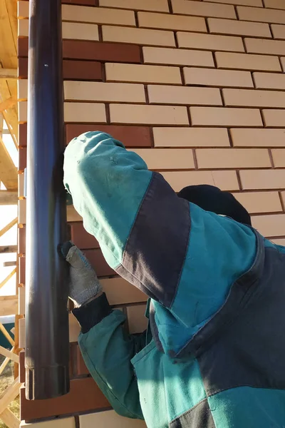 To the house bricked with a front brick, the worker fastens pipes for drainage. the photo shows the wall, brown pipes and the worker\'s hands 2020.