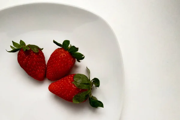 Fraises fraîches maison sur une assiette en porcelaine blanche, trois fraises juteuses sur une assiette, assiette brillante et blanche. — Photo