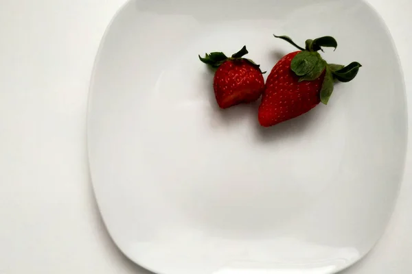 Deux fraises sur une assiette, une fraise mordue, gros plan, baies sur une assiette blanche brillante, place pour une inscription. — Photo