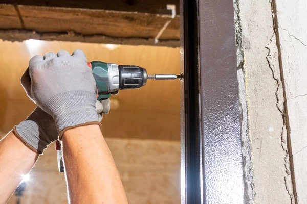 The carpenter drills holes in the wall and the metal door frame with a drill, later screws the screws and fastens the door frame to the wall.2020