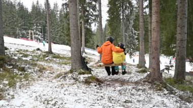 Bir ebeveyn ve çocuğun ağaçlar arasında asılı bir kırsal salıncakta bir anı paylaştığı yürek ısıtan bir sahne. Karlı bir manzara ve uzak bir kayak yamacı Kış Harikalar Diyarı atmosferine katkı sağlar.