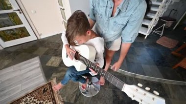 Genç bir çocuk bir taburede oturuyor, bakımlı bir yetişkinin rehberliğinde beyaz bir gitarı tıngırdatmaya odaklanmış, şık bir ev dekoruyla çevrili..