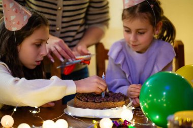 İki genç kız çikolatalı pastayı şekerle süslemeye odaklanırken balonlar ve peri ışıkları sıcak, davetkar bir odada neşeli bir atmosfer yaratıyor.