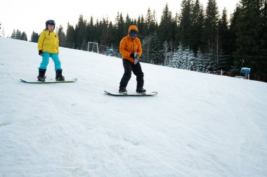 İki snowboardcu açık bir gökyüzünün altında, yüksek çam ağaçlarıyla çevrili karlı bir tepeden aşağı süzülüyor, kış sporları macerası sırasında kahkaha ve neşe paylaşıyorlar..