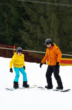Bir çocuk, kar yamacındaki bir eğitmenden, yemyeşil ağaçların ve açık mavi gökyüzünün arasında parlayan güneş sporu becerilerini öğrenir..