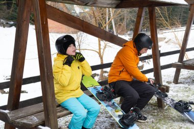 Parlak giyimli çocuklar bir bankta oturup snowboard yapmaya hazırlanıyorlar. Zevkle sohbet ediyorlar, yamaçlardaki eğlenceli maceralarına hazırlanırken karlı manzaranın tadını çıkarıyorlar..