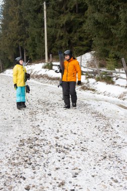 Kış kıyafetleri giymiş iki hevesli snowboardcu kar kaplı bir dağ yolunda kar sporlarının maceralı bir gününe hazırlanarak sohbet ediyorlar..