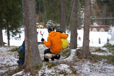 İki kayakçı, dinlenmek için etrafı karla kaplı bir salıncakta mola veriyor. Arkaplanda kar meraklıları var..