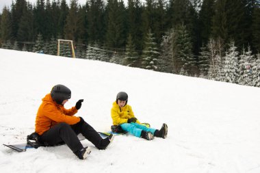 Parlak ceketli snowboardcular karda oturup gülüyorlar ve hikayelerini paylaşıyorlar. Dağlarda sakin bir kış gününün tadını çıkarıyorlar..