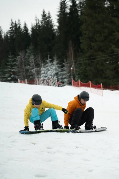 İki arkadaş karlı bir yamaçta oturmuş birlikte snowboard yapmaya hazırlanıyorlar. Parlak ceketleri beyaz manzaralarla tezat oluşturarak canlı bir kış ortamı yaratıyor..