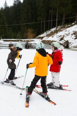 Üç çocuk uzun çam ağaçlarıyla çevrili, karlı bir yamaçta birlikte kayak yapıyor. Kask takıp renkli ceketler giyiyorlar. Dağlarda kış eğlencesinin tadını çıkarıyorlar..