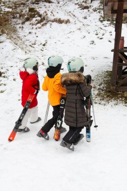 Canlı kayak kıyafeti giymiş üç heyecanlı çocuk, kış boyunca karlı bir yerde kayak yapmaya hazır bir şekilde asansöre doğru yürüyorlar..