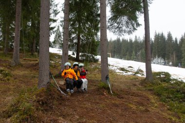 Neşeli çocuklar güçlü iplerden sallanırken gülerler. Parlak ceketleri, yaklaşan kışın kar ima ettiği sakin orman zeminiyle çelişir..