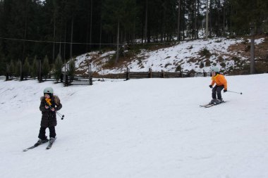 Çocuklar, yemyeşil çamlar ve parlak gökyüzü ile çevrili kayak yaparken karlı yamaçları keşfeder, eğlence ve macera dolu mükemmel bir kış atmosferi sergilerler.