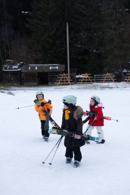 Üç çocuk ellerinde kayak ekipmanlarıyla karlı bir arazide coşkuyla yürüyorlar. Parlak kış gökyüzünün altında kayak yapmaya hazırlanıyorlar..