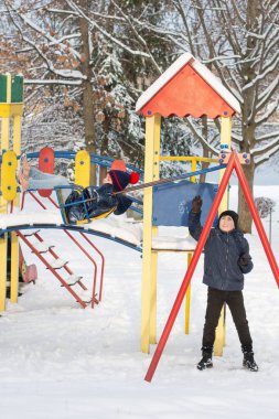 İki çocuk karla kaplı bir oyun alanında kış gününün tadını çıkarıyor. Bir çocuk tahterevallide sallanırken diğeri sabırla sırasını bekliyor. Sahne kış cazibesiyle dolu..