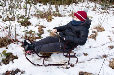 Genç bir insan, karlı ve ağaçlık bir alanda küçük, koyu renkli bir kızağın üzerinde oturur. Küçük bir dalları var, görünüşe göre huzurlu bir kış gününün tadını çıkarıyorlar. Kızak bir iple çekiliyor..