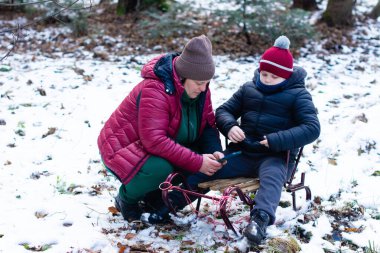 Bir yetişkin ve küçük bir çocuk kış ormanı ortamında karın üzerinde oturuyorlar. Birlikte küçük bir ahşap kızağın yanındaki bir cep telefonuna bakıyorlar..