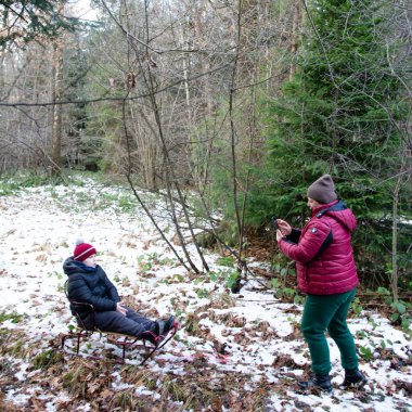 Küçük bir çocuk, karlı bir ormandaki küçük bir kızakta kış gününün tadını çıkarıyor. Yakınlarda bir yetişkin duruyor, bir telefonla anı yakalıyor..