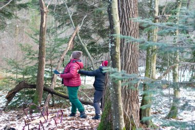 Karlı, ağaçlık bir alanda, iki birey araziyi araştırıyor. Bir kişi tablet tutuyor, muhtemelen harita çıkarmak ya da veri toplamak için. Yakınlarda kırık bir ağaç dalı uzanıyor ve karda bir kızak dinleniyor..