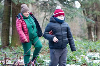 Bir yetişkin ve bir genç, karla kaplanmış ağaçlık bir yolda yürüyor. Arkalarında küçük bir kızak çekerler, açık havada kış gününün tadını çıkarırlar..