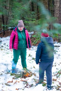 İki figür, bir yetişkin ve bir çocuk, bir kış ormanı ortamında küçük bir ateşin yanında duruyor. Kar zemini kaplıyor ve her yer yemyeşil ağaçlarla çevrili. Sıcaklığın tadını çıkarıyor gibi görünüyorlar..