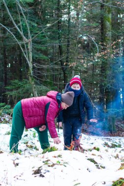 Kırmızı ceketli bir insan, karlı ve ağaçlık bir alanda küçük bir ateş yakarak bir çocuğa yardım eder. Sahne huzurlu ve kışlık..