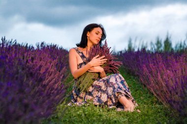 Gün batımında canlı bir lavanta tarlasında oturan bir kadının huzurlu görüntüsü. Büyük bir demet lavanta tutuyor, ifadesi huzurlu ve memnun. Ortam huzurlu ve huzurlu..