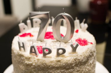 Kutlama pastası, kremalı beyaz ve gümüş mumlarla süslenmiş 70 tane ve daha küçük mum şeklinde Happy kelimesini oluşturuyor. Kırmızı aksanlar şenlikli bir dokunuş ekler.