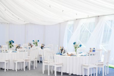Saf perdeleri, zarif dekore edilmiş masaları, beyaz sandalyeleri ve mavi çiçekleri olan el değmemiş beyaz bir parti çadırı. Sahne, sofistike ve kutlama hissi uyandırıyor..