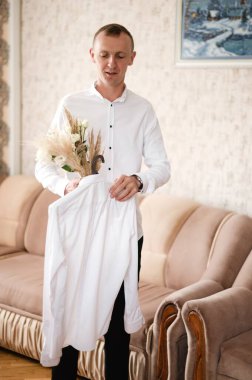 Beyaz gömlekli bir adam temiz bir gömlek ve bir buket kuru çiçek ve beyaz güller tutuyor. Oturma odasında duruyor, bej bir kanepenin yanında, hazırlanıyor..