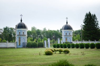 Görüntü, düzgün kesilmiş çalılar ve iyi korunmuş bir çimenlikle çevrili süslemeli bir kapının yan tarafında ikiz açık mavi kubbeli binaların bulunduğu sakin bir manzara gösteriyor..