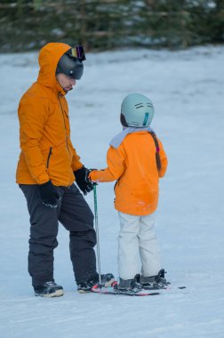 Yetenekli bir kayak hocası, her ikisi de parlak turuncu ceketler giymiş, karlı bir yamaçta genç bir çocuğa rehberlik eder. Kışın harikalar diyarında öğrenme ve heyecan anlarını paylaşıyorlar..