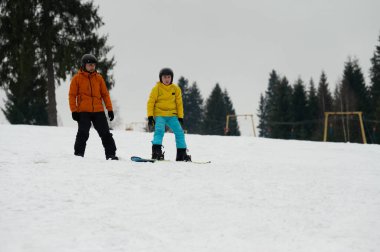 İki snowboardcu karlı bir yamaçta süzülmeye hazır bekliyor. Atmosfer kıtır kıtır ve yüksek çam ağaçları etraflarını sararak mükemmel bir kış ortamı oluşturuyor..