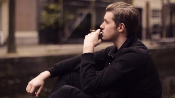 アムステルダムでの男のマリファナ アムステルダムの路上で大麻のジョイントを吸って少年 遅いミズーリ 観光川背景に少年喫煙雑草しようとコーヒー ショップの近くの大麻 — ストック動画