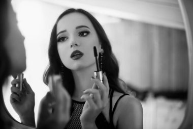 Güzel bir genç kadın kendini bir makyaj yapıyor. Siyah ve beyaz ışık ayna önünde maskara kullanarak akşam makyaj cerly saçlı kız. Clouse kırmızı dudaklar ile kız.