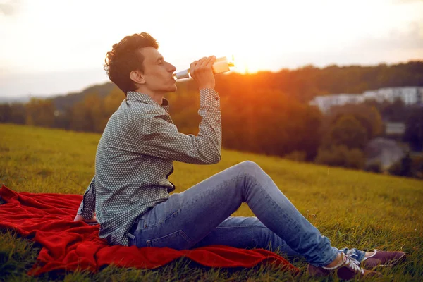 Man portrait  on sunset. Man sitting at the park on a summer day and drinking beer. Side view of handsome young man drinking cider. Beautiful view of the city