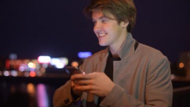 Geceleri dışarıda gülümseyen yakışıklı genç bir adam. Adam mesajlaşırken gece akıllı telefondaki uygulamayı kullanıyor. Akıllı telefon kullanan yakışıklı genç bir öğrenci gülümseyerek mutlu bir şekilde takım elbise giyiyor.